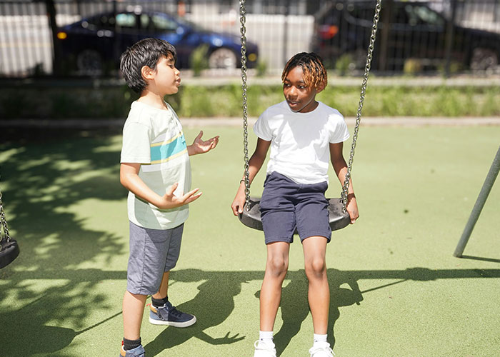 Two children in a playground, one sitting on a swing and the other standing, discussing overused slang words to banish. Two children in a playground, one sitting on a swing and the other standing, discussing overused slang words to banish.