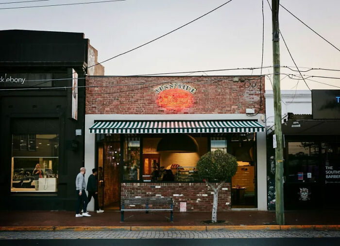 Brick pizzeria exterior with neon sign and striped awning, known for owner&rsquo;s viral salty replies to online reviews.