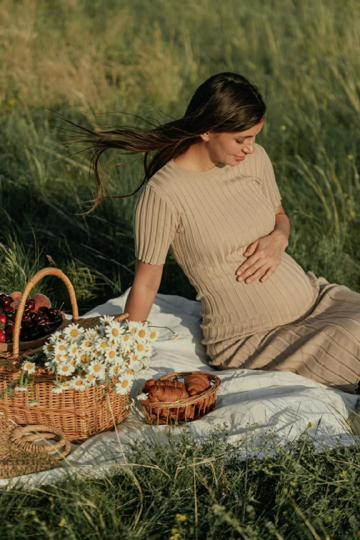 Pregnant woman on picnic blanket in meadow holding belly with baskets of daisies and croissants, maternity photoshoot ideas