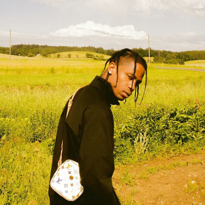 Young man with braided hair wearing a black jacket and designer bag in a field, representing movie set workers and diva stars.