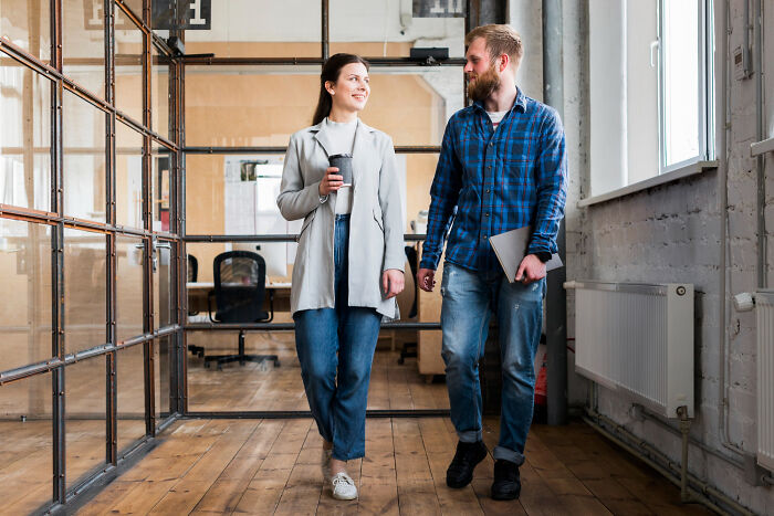 Two coworkers walking and chatting in a modern office with glass walls, illustrating bizarre office rules frustration.
