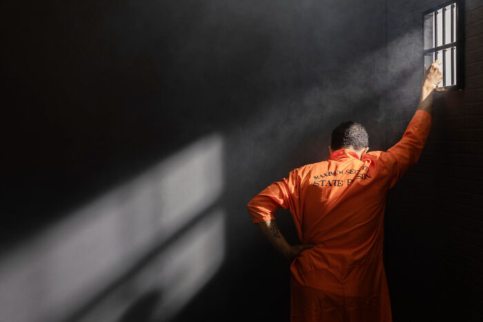Prisoner in orange jumpsuit leans against barred window in dark cell, illustrating misconceptions about life in prison.