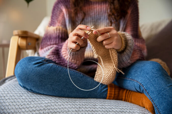 Person sitting cross-legged on a couch knitting a beige fabric, illustrating bizarre office rules boredom concept.