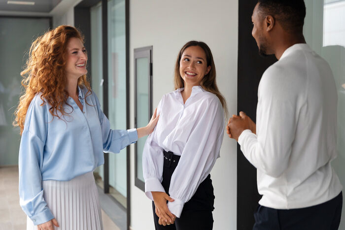 Three diverse coworkers smiling and chatting in an office hallway, illustrating bizarre office rules causing frustration.