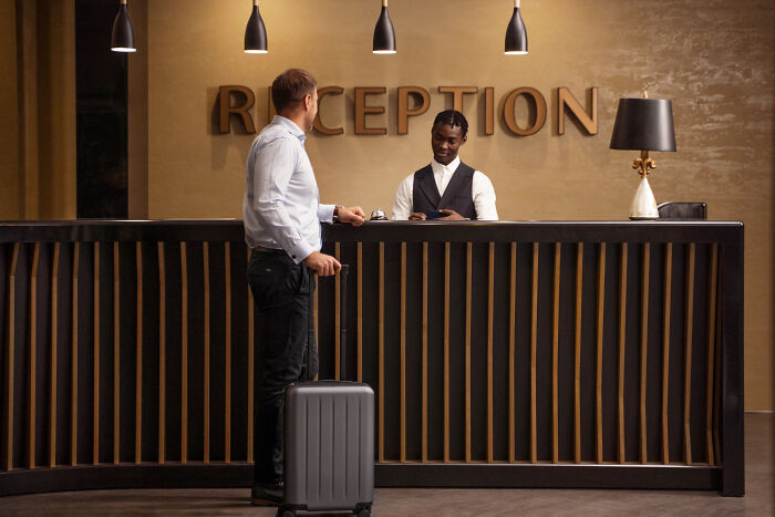 Man with suitcase at hotel reception desk talking to receptionist in a moment so ridiculous people thought they were being pranked
