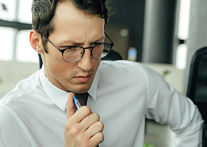 Young man wearing glasses and white shirt, thoughtfully holding a pen, illustrating neurodivergent habits in focus.
