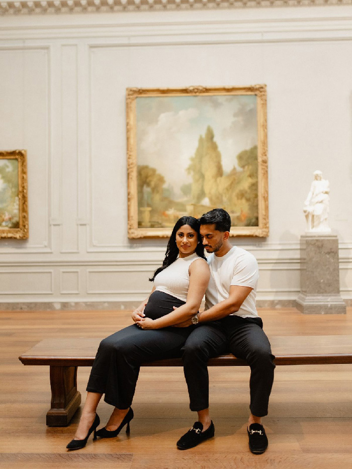 Pregnant couple seated on museum bench, woman in white top and black pants, partner holding belly, maternity photoshoot