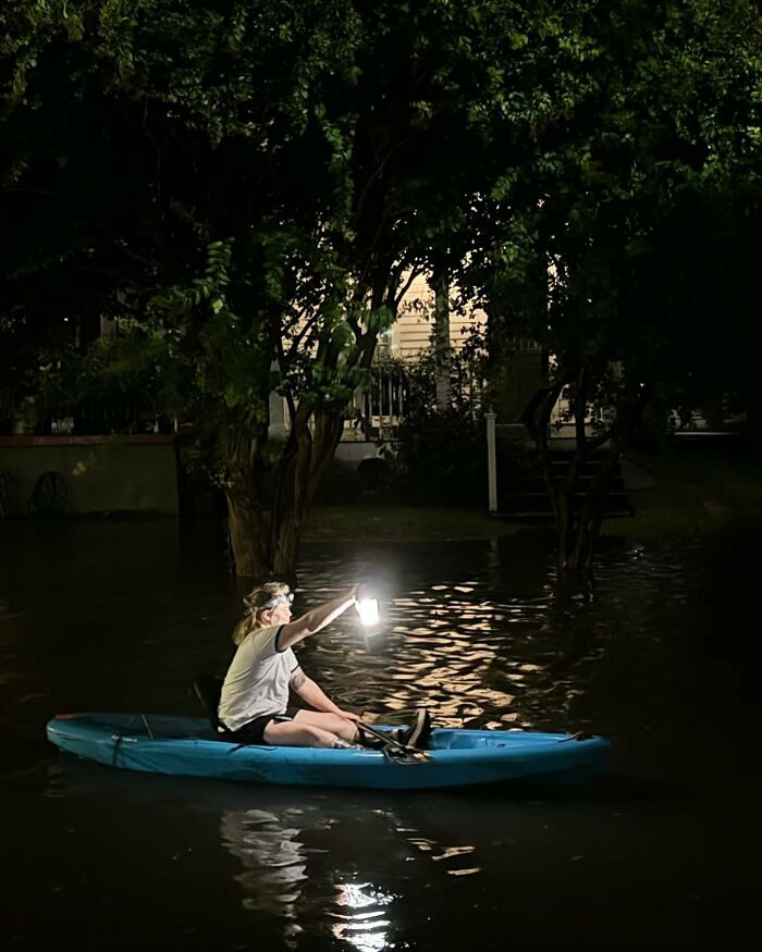 Person in a kayak holding a light while navigating flooded streets surrounded by trees at night, a weird street finding.