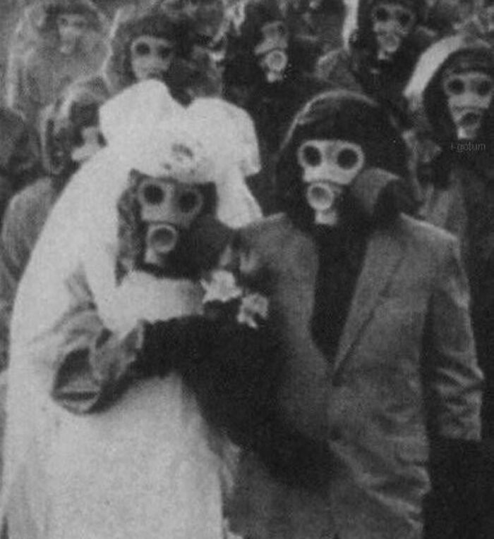 Couple in wedding attire wearing gas masks among a crowd, an old black and white creepy and odd photo.