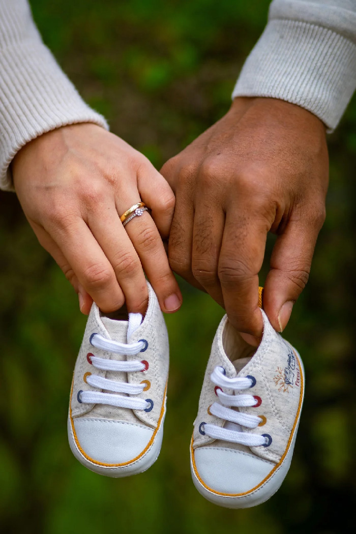 Couple hands holding baby sneakers for maternity photoshoot, close-up of rings and tiny shoes against soft green background