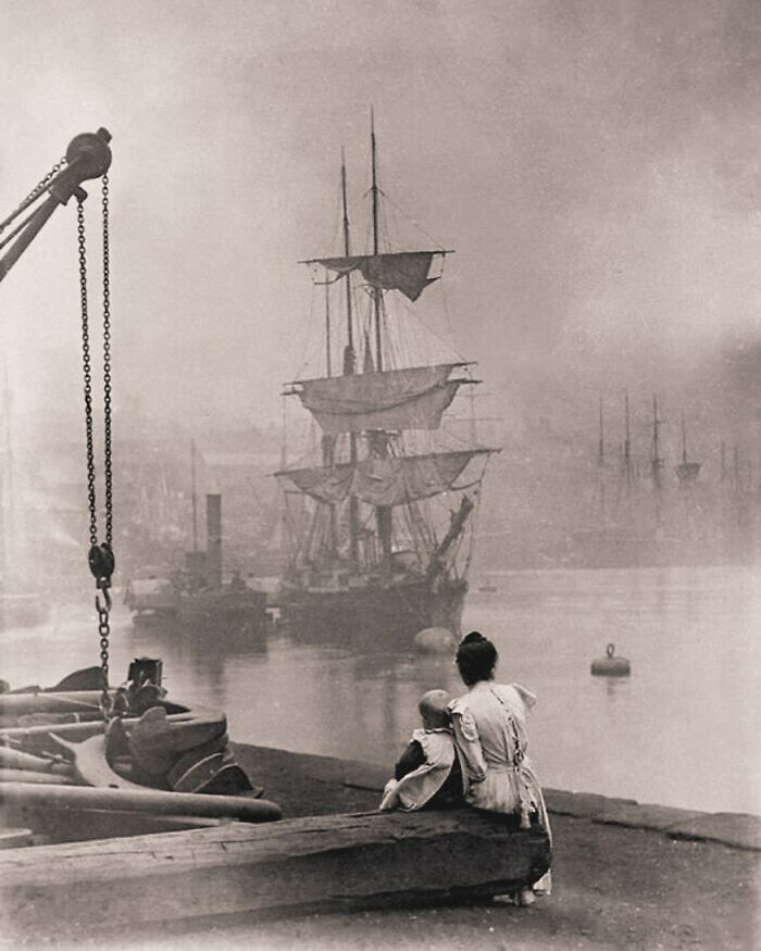 Mother and child sitting by a foggy harbor with old sailing ships, capturing a historical moment in humanity's past.