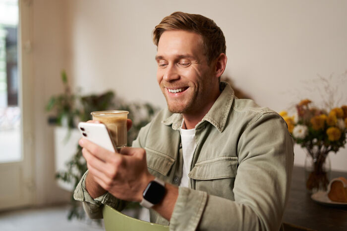 Smiling man holding coffee and phone in cozy room, enjoying a satisfying moment away from problems.