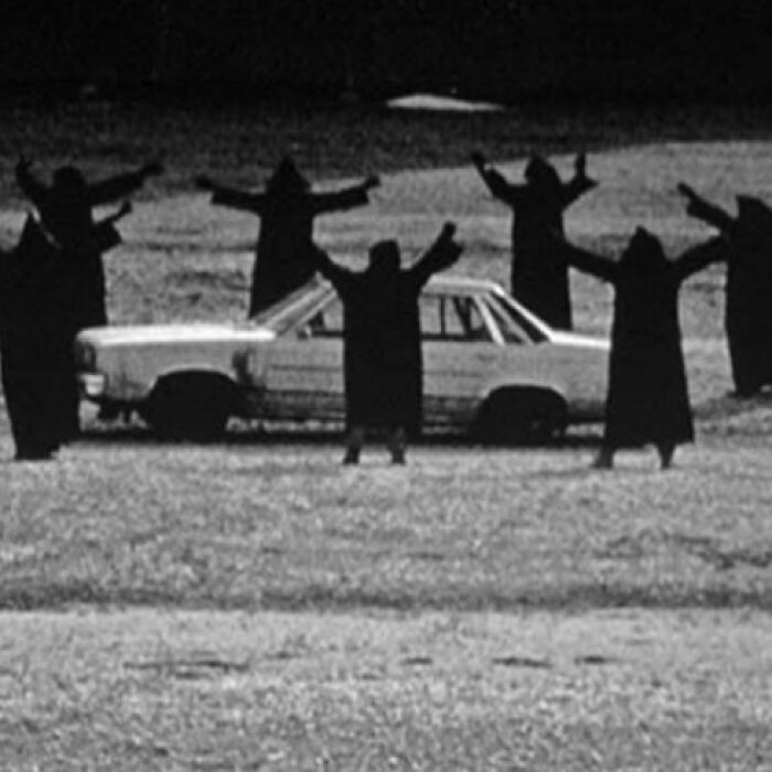 Group of six robed figures with arms raised around an old car in an odd and creepy black and white scene.