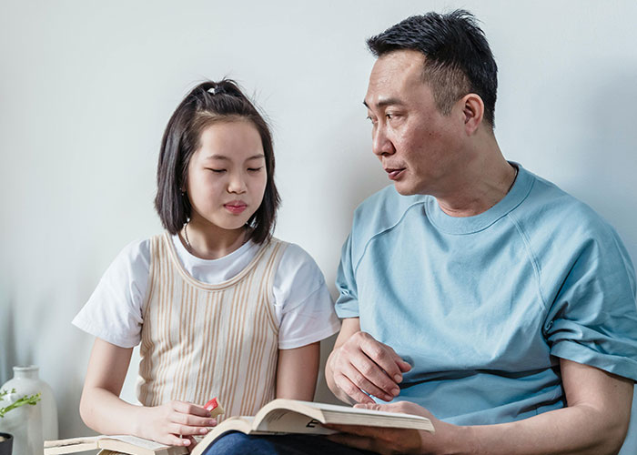 Asian man and young girl reading a book together, depicting emotions of discovering he is not the biological parent.