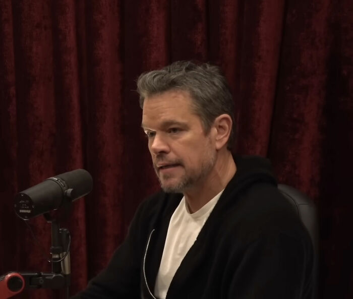 Matt Damon in a podcast setting, speaking into a microphone with a red curtain backdrop, related to cancel culture comments.