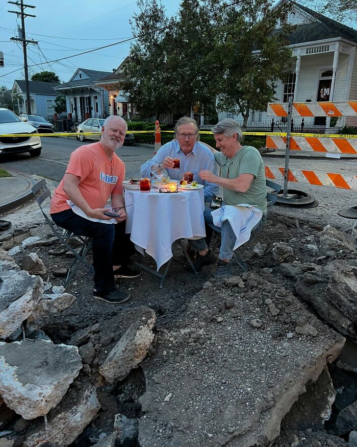 Three men dining at a table set up on a broken street surrounded by rubble, a weird thing found in the streets.