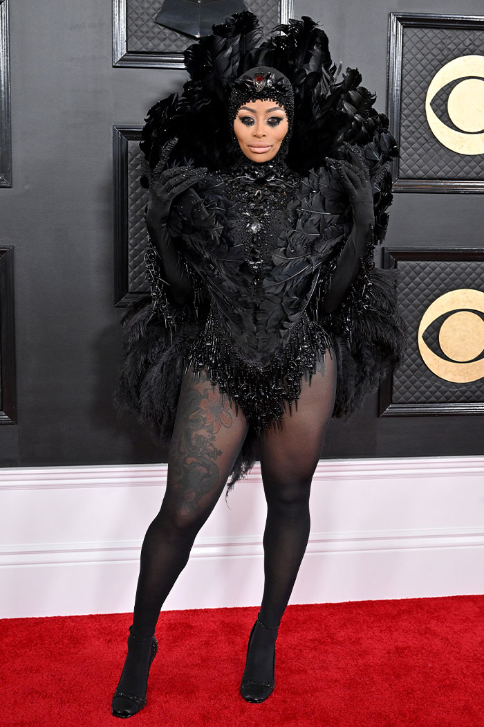 Grammy red carpet look featuring a bold black feather and beaded outfit with dramatic headpiece and sheer tights.