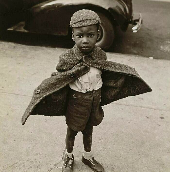 Young boy wearing oversized coat and cap standing on street, capturing sad and beautiful moments in history of humanity.