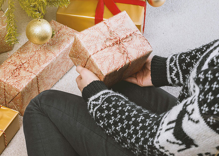 Person holding wrapped gift sitting by Christmas tree with ungrateful feelings about husband's gifts debated online