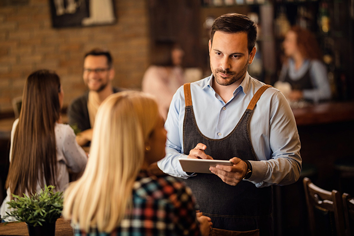 Waiter in apron taking order at restaurant table while customers talk in the background.