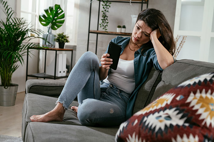 Woman sitting on couch looking upset at phone, reflecting on karma after affair partner leaves her alone. Woman sitting on couch looking upset at phone, reflecting on karma after affair partner leaves her alone.