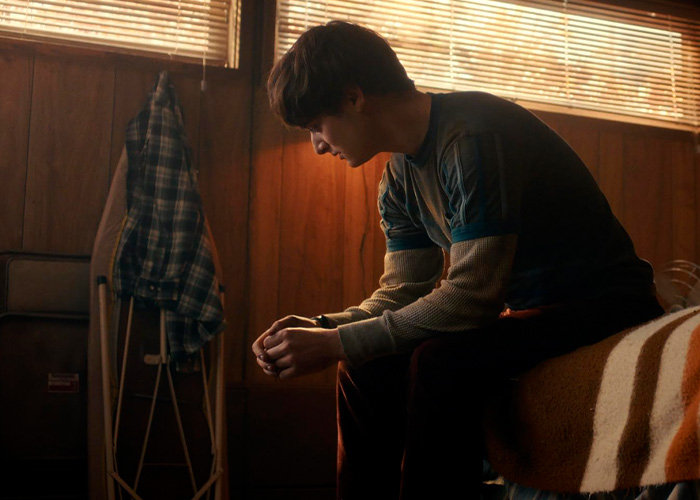 Noah Schnapp as Will Byers sitting on a bed in a dimly lit room with wooden walls and an ironing board nearby Noah Schnapp as Will Byers sitting on a bed in a dimly lit room with wooden walls and an ironing board nearby