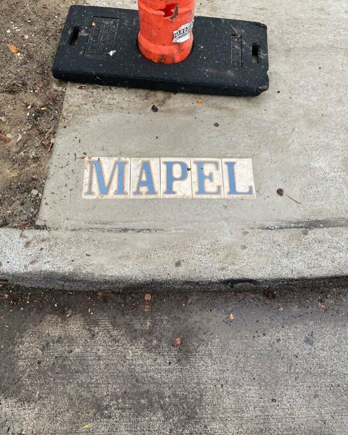 Tiles spelling MAPEL on a street curb near an orange construction cone, showing weird street finds.