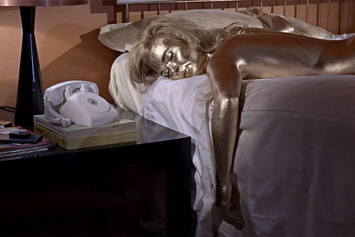 Gold-painted model lying face down on bed, arm dangling off edge, bedside table with white rotary phone, photoshoot