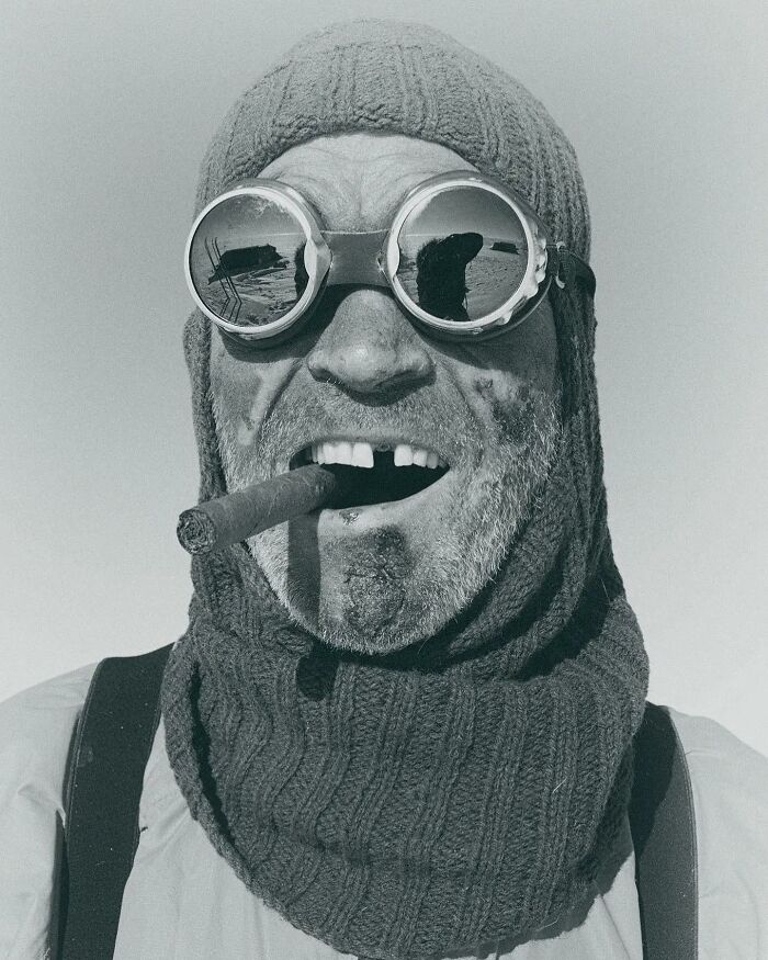 Man wearing knitted hat and reflective goggles, smoking a cigar, captured in a historic photograph showing sad and beautiful humanity.