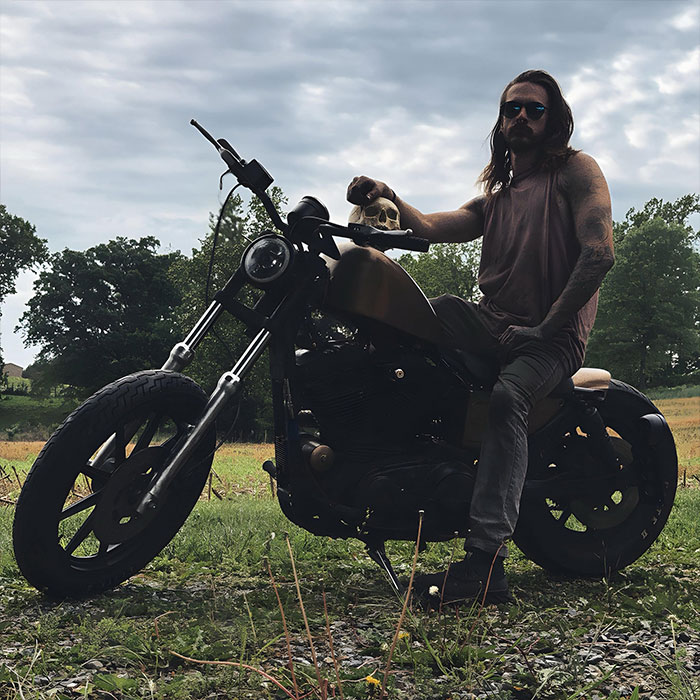 Man sitting on a motorcycle holding a skull outdoors with trees in the background, suggesting a chilling discovery. Man sitting on a motorcycle holding a skull outdoors with trees in the background, suggesting a chilling discovery.