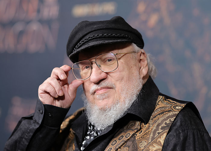 George RR Martin wearing a cap and glasses, posing at a promotional event for A Knight of the Seven Kingdoms release.
