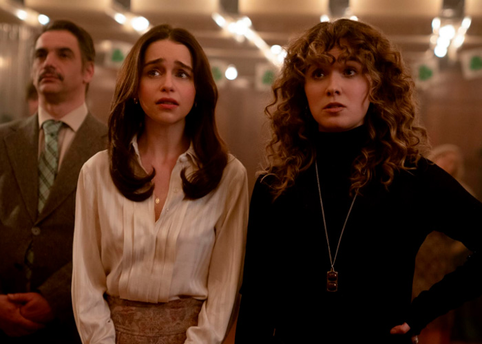 Emilia Clarke in a tense scene wearing a white blouse, standing next to a woman with curly hair in a black top indoors.