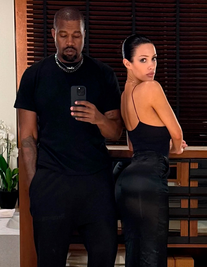 Bianca Censori wearing sleek black pants posing confidently next to a man in a black shirt taking a mirror selfie.