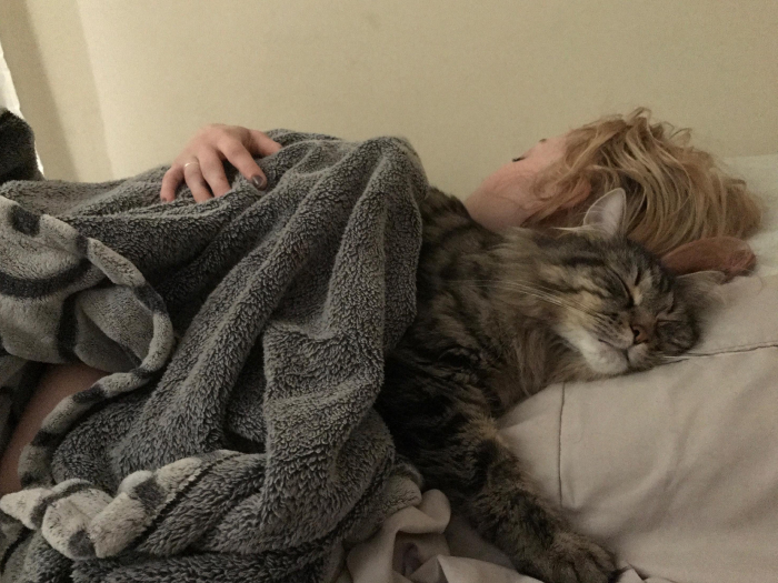 Woman sleeping under a gray blanket with a striped cat resting beside her, illustrating sleep needs in women.