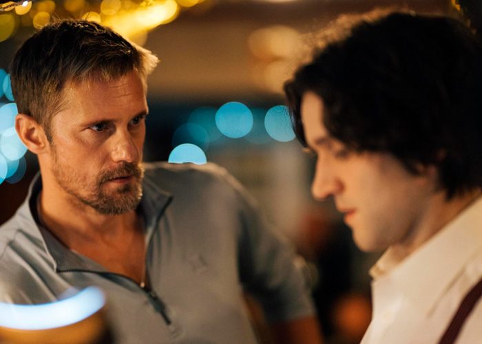 Alexander Skarsg&aring;rd in a tense conversation with another man in a dimly lit setting, focusing on orientation discussion.