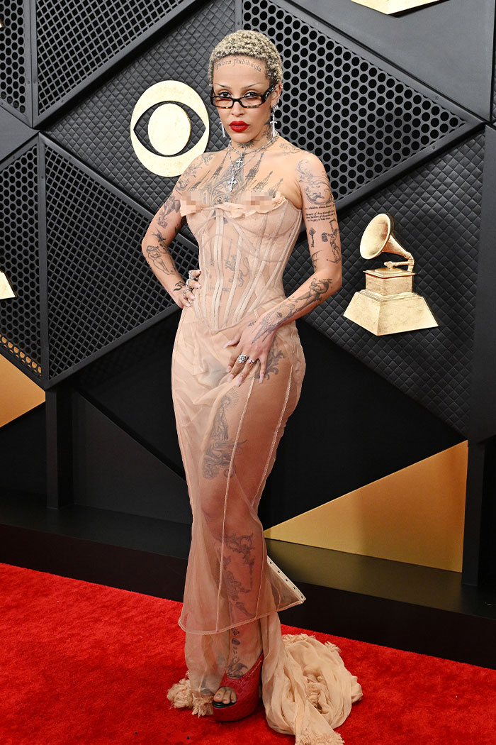 Model with tattoos in a sheer beige dress and red platform heels posing on the red carpet at a wild Grammys look event.