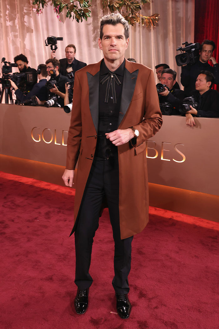 Man in brown coat and black outfit posing on the red carpet at 2026 Golden Globes fashion event with photographers behind him