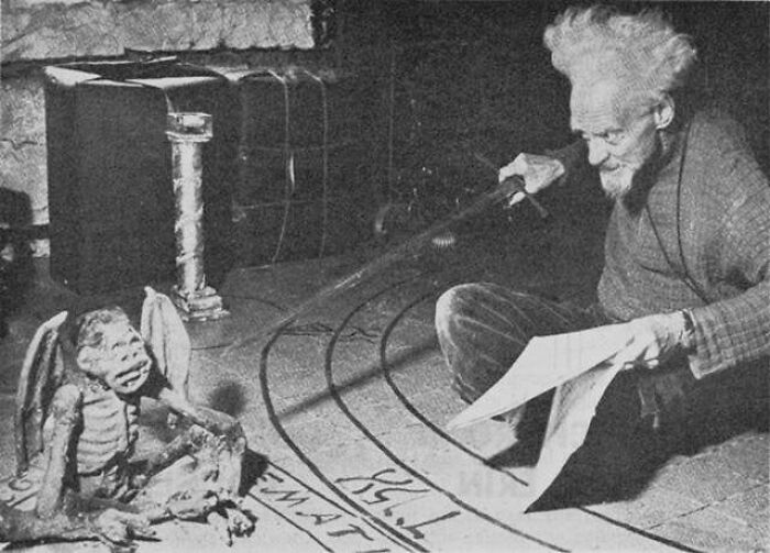 Old black and white creepy photo of a man interacting with a strange winged creature in a dimly lit room.