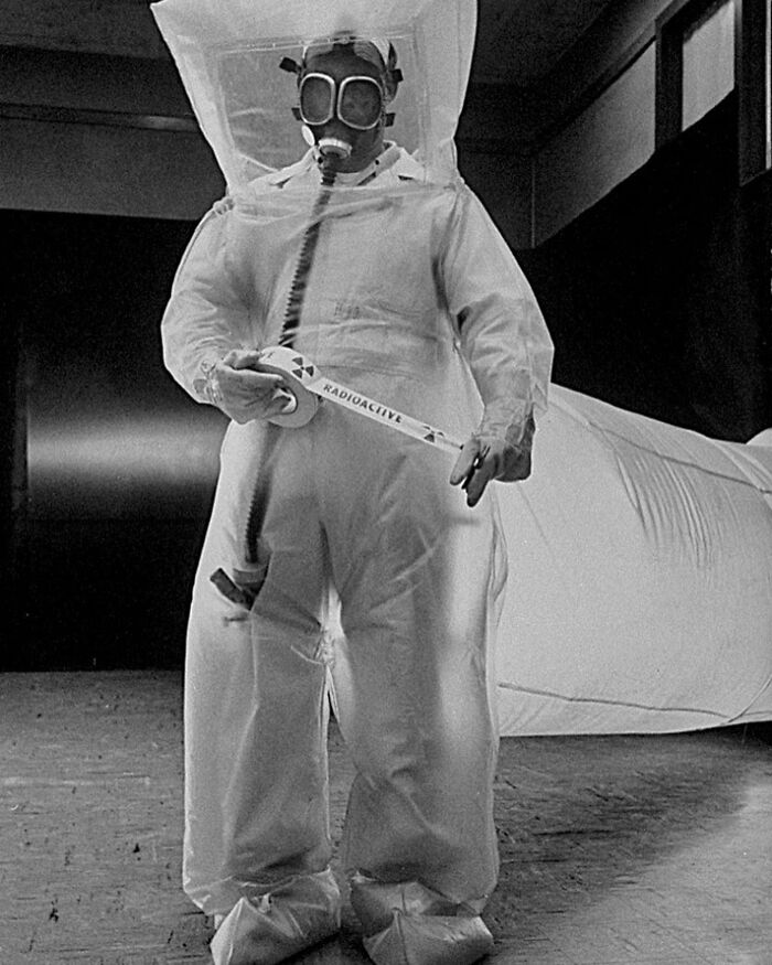 Person in an old black and white photo wearing a protective suit and gas mask holding radioactive warning tape in a creepy setting.