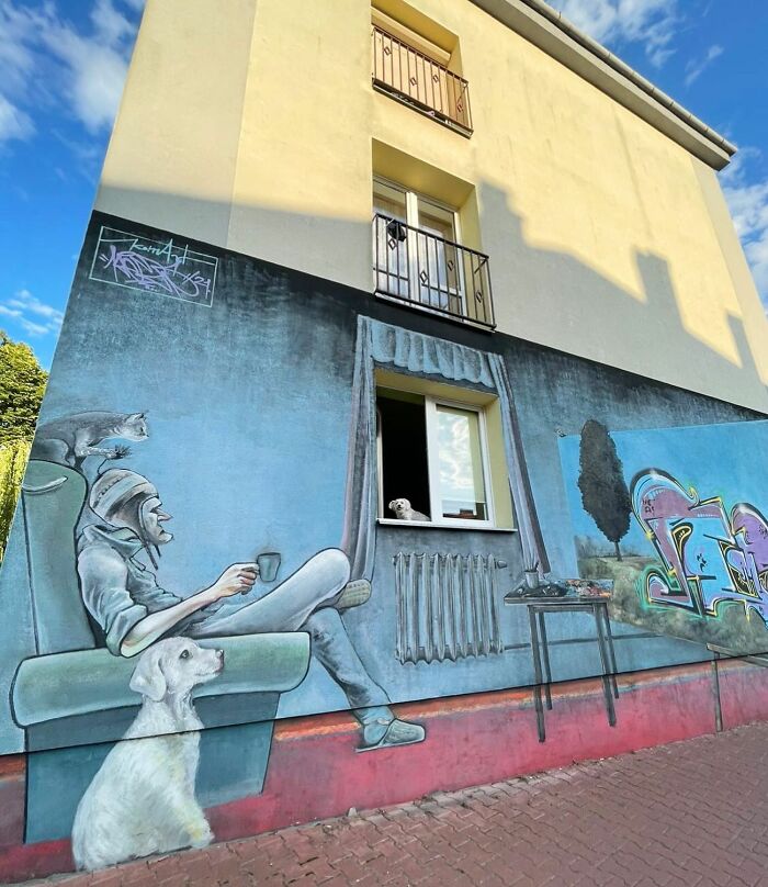 Adorable dog peeking out of a real window set within a mural featuring a person and painted dog relaxing indoors.