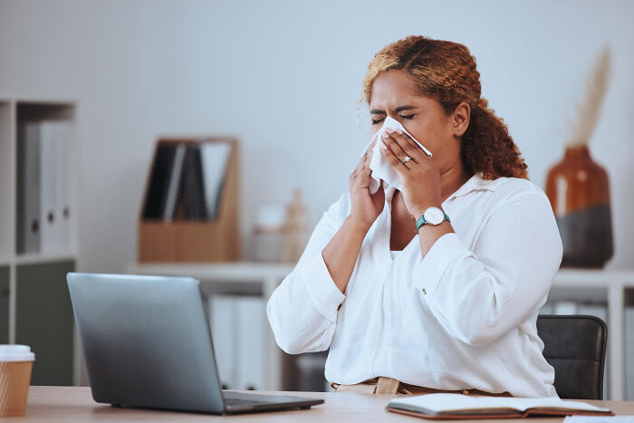 Woman at office desk blowing nose in tissue, illustrating bizarre office rules causing frustration and discomfort.