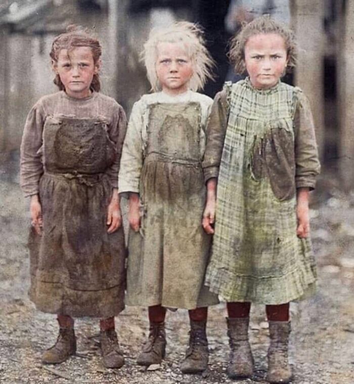 Three young girls in worn, dirty clothing standing outside, reflecting the sad history of humanity captured in a photograph.