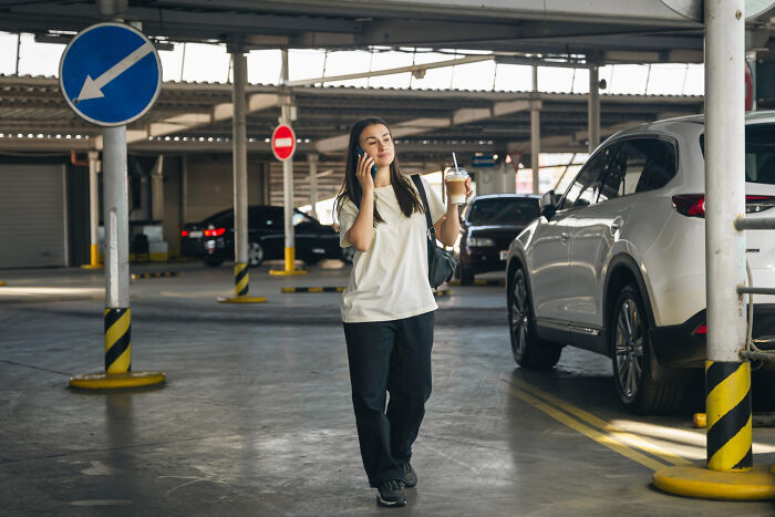 Young woman holding coffee and talking on phone in parking garage, illustrating bizarre office rules causing frustration.