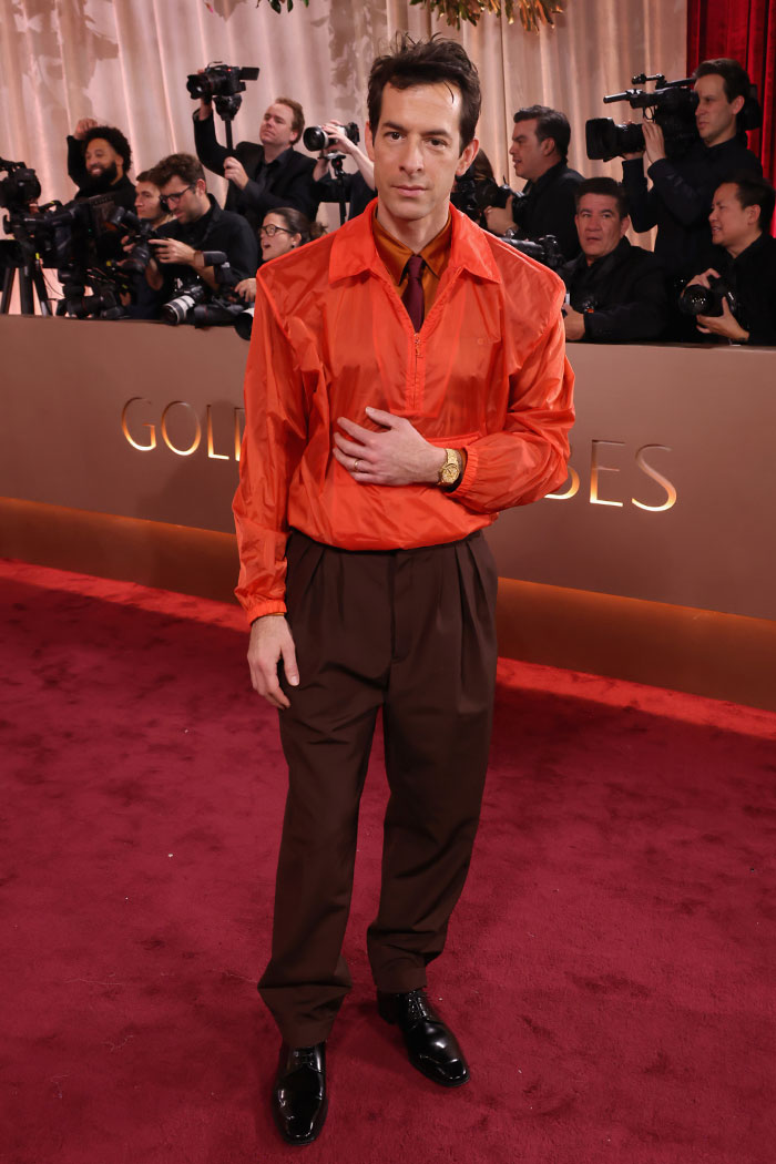 Man in an orange jacket and brown pants posing on the red carpet amid Golden Globes fashion disasters and stylist controversies.