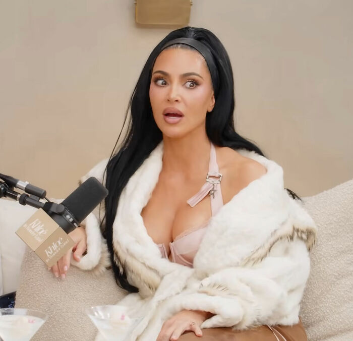 Kim Kardashian speaking into a microphone, wearing a white fur coat and looking surprised during an interview.