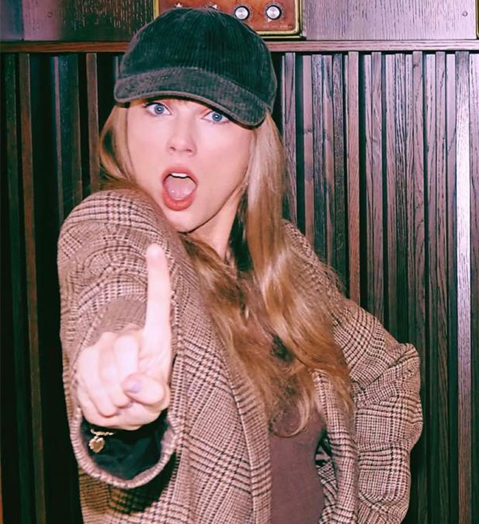 Taylor Swift wearing a cap and plaid jacket, making a bold gesture with an intense facial expression indoors.