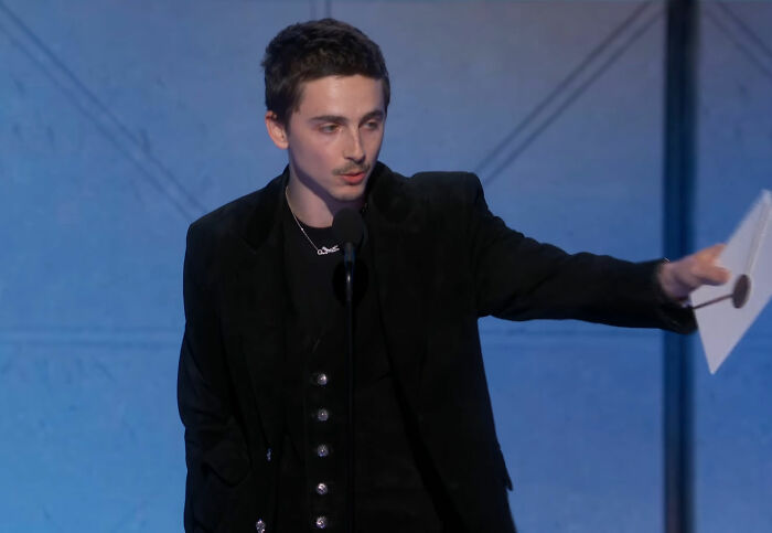 Actor Timoth&eacute;e Chalamet in a black suit speaking at a podium during the Golden Globes, sparking media buzz about snubs.