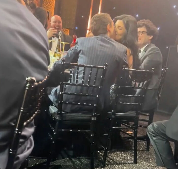 Timoth&eacute;e Chalamet at critics&rsquo; choice awards, sitting and standing with guests in a dimly lit event setting.