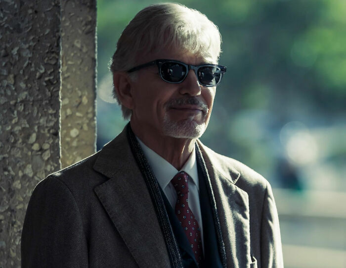 Older man in sunglasses and suit standing by a pillar, representing movie set workers and diva stars in film industry.