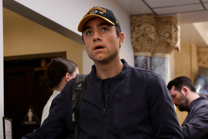 Actor in a scene from The Pitt Season 2 Episode 1, wearing a cap and backpack inside a building.
